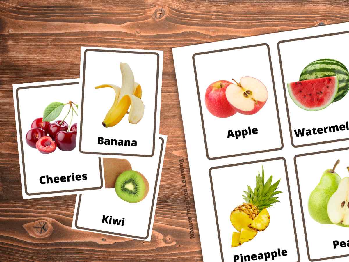 printed out flashcards with apple, watermelon, pineapple, and pear next to a banana, kiwi, and cherries flashcards all on a wooden background.