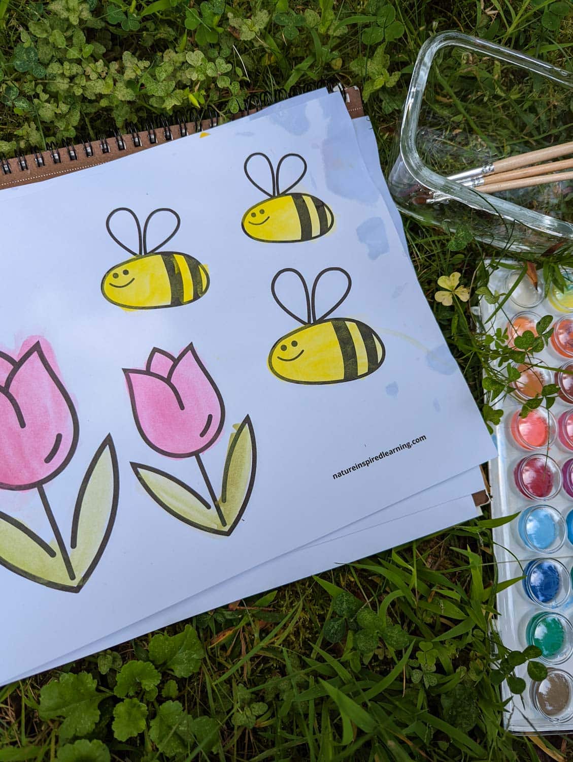 coloring page with three smiling bees with two tulips painted using watercolor paint outside in the grass with a glass with paint brushes and a watercolor paint set.