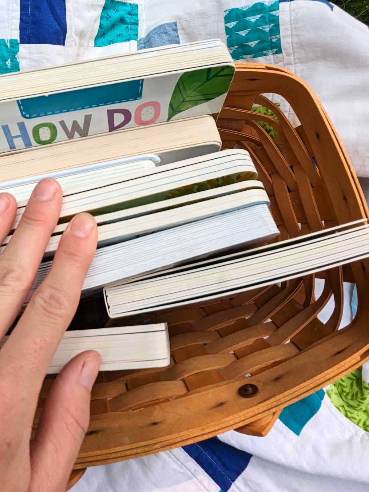 collection of board books in a woven basket on a colorful quilt with a hand on top of them on the left side.