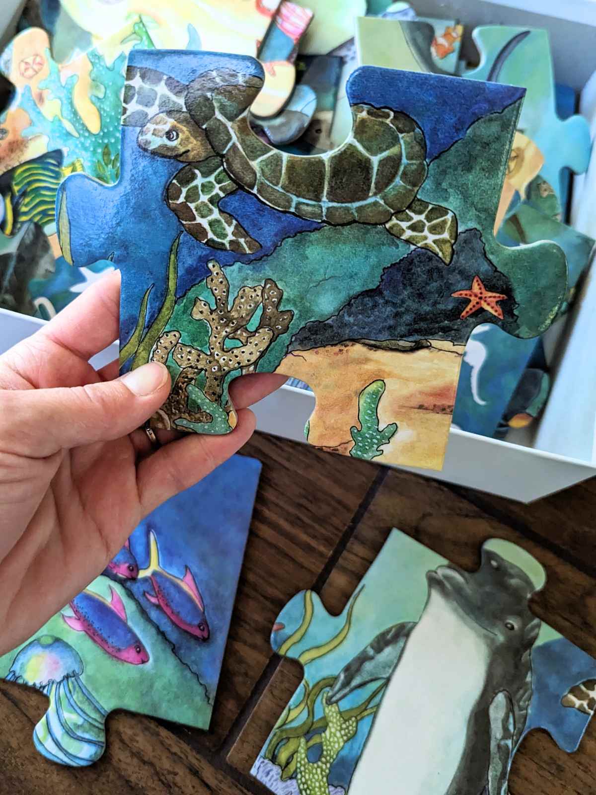 hand holding a large puzzle piece with a sea turtle, coral, and starfish. White box on the floor with puzzle pieces along with two ocean themed puzzle pieces on a wooden floor.