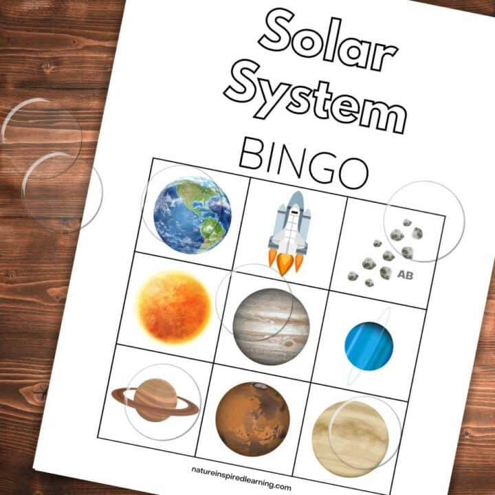 Bingo card with planets, a space ship, and asteroids slanted on a wooden background with clear plastic bingo chips on top.