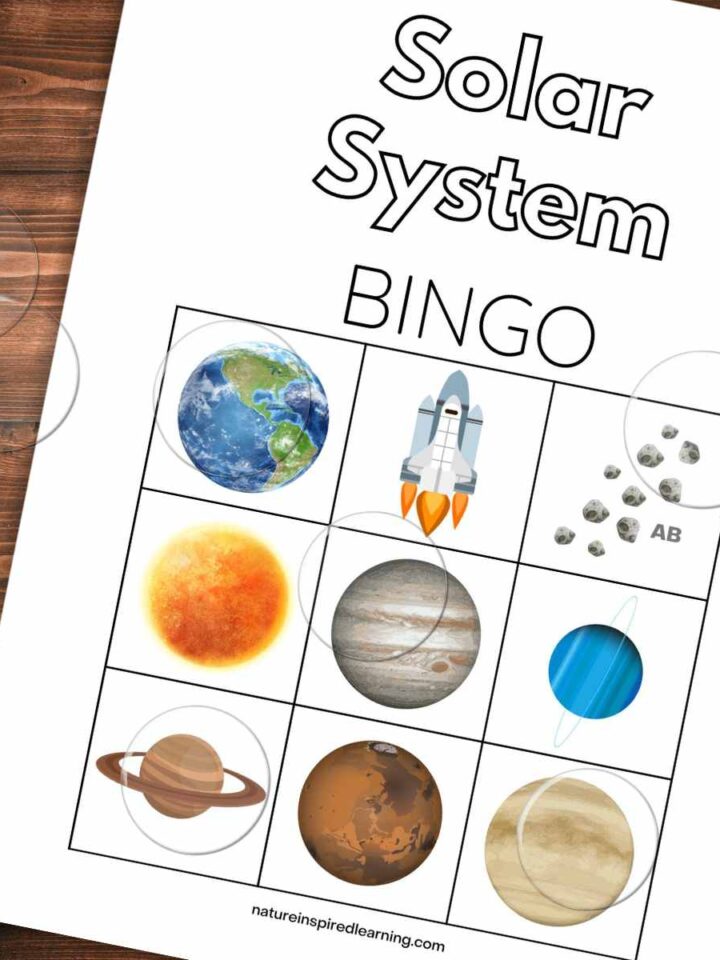 Bingo card with planets, a space ship, and asteroids slanted on a wooden background with clear plastic bingo chips on top.
