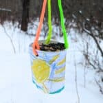 DIY bird feeder made out of a cup with a bright yellow sun drawn on it hanging from colorful pipe cleaners outside in the winter. Snow and bare branches in the background.