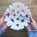 Hands holding a handmade snowflake using a coffee filter over a wooden surface. Light blue sweater bottom corners.