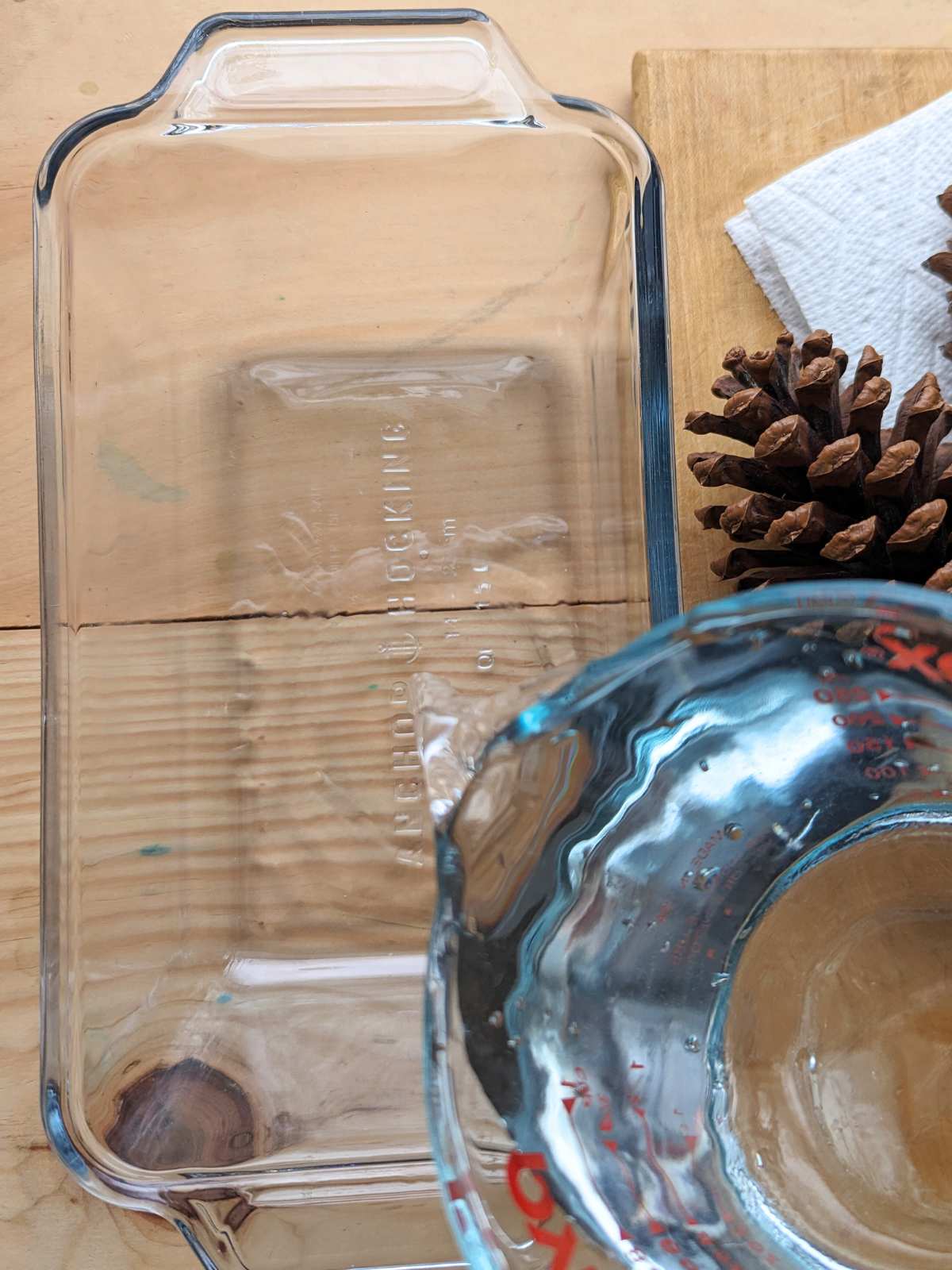 Glass measuring cup pouring water into a glass dish on a wooden table with a pinecone and paper towel on the right.