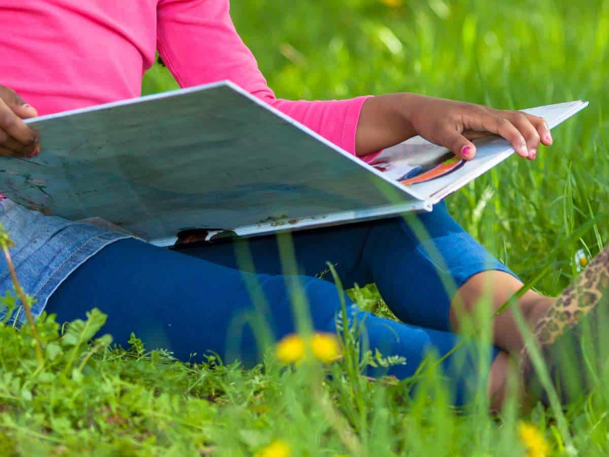 Child wearing pink shirt, jean shorts, and blue leggings with leopard print shoes sitting in the grass with dandelions reading a picture book on their lap.